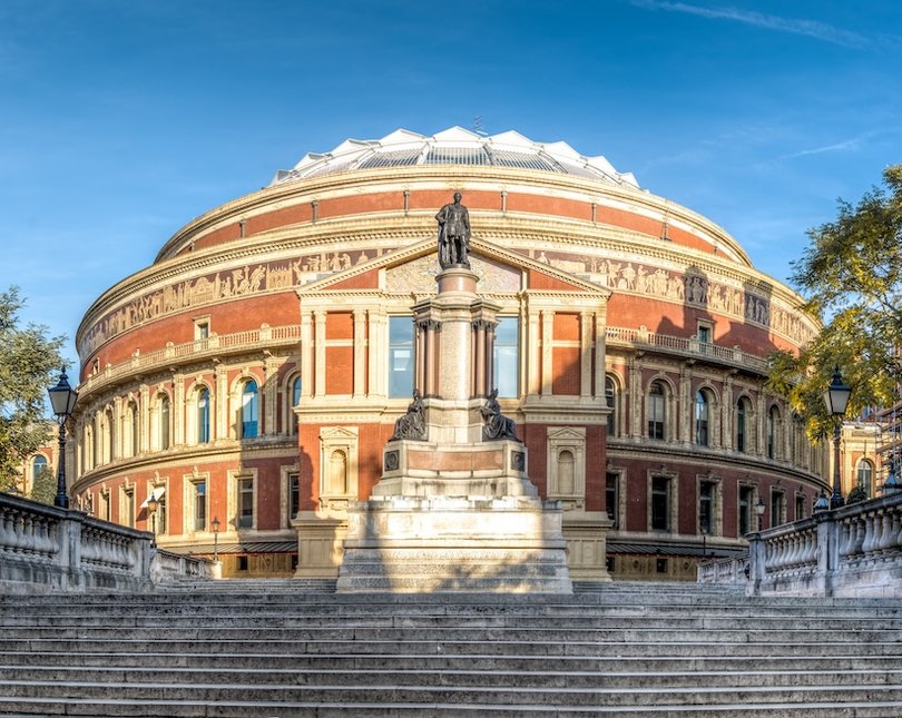 Photo of the Royal Albert Hall (a large round building) in the sun with a statue in front