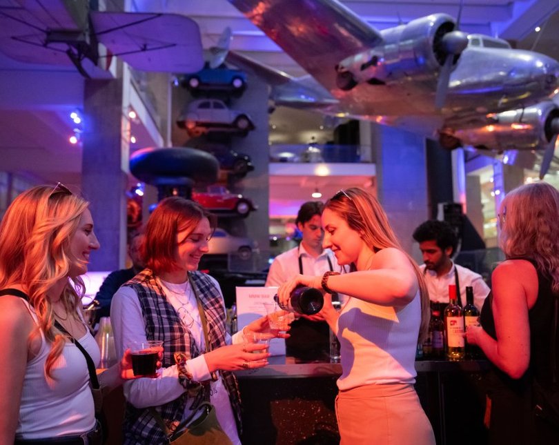 Group of girls pouring drinks inside a room with a plane hanging above them (at the Science Museum)