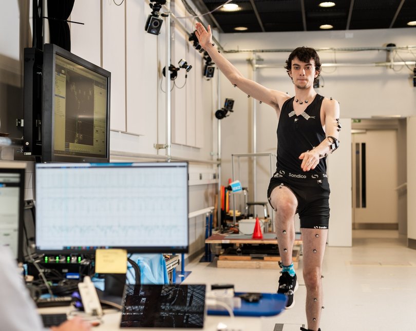 Man covered in small sensors lifting one harm and one knee, with another man looking at data on a computer