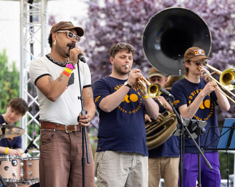 Singing wearing sunglasses on stage with two musicians playing the trumpet and trombone next to him