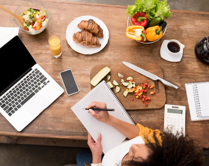 A desk covered in food and a woman taking notes next to a laptop