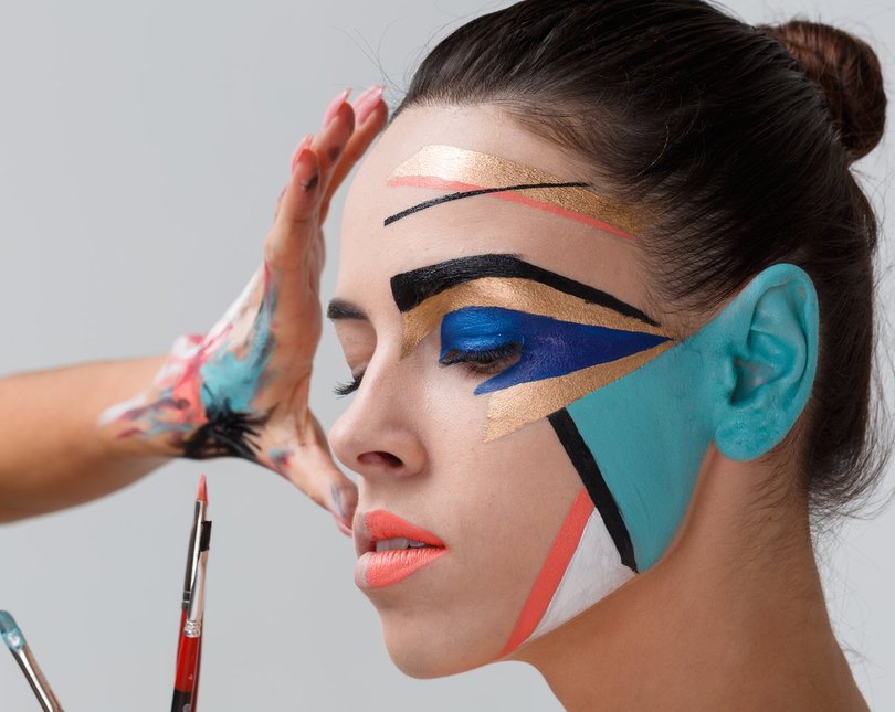 A woman closes her eyes while geometric patterns are applied in bright face paints across her cheek and eye